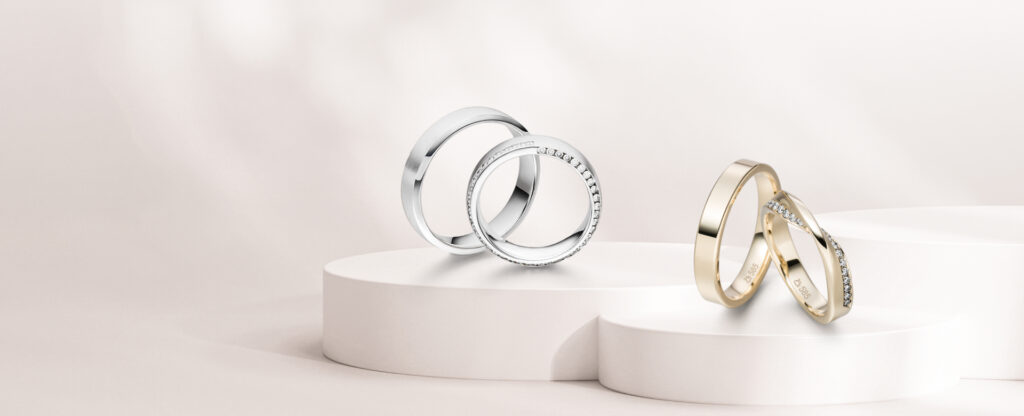 Ein weißes und ein goldfarbenes Trauringpaar auf weißen Podesten, je ein Ring mit Brillanten